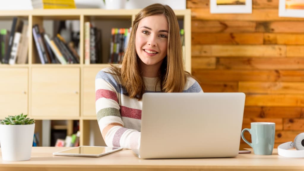 A person sits at a desk with a laptop, tablet, and mug, surrounded by a small plant, smiling at the camera while designing a mental health website. Bookshelves and a wood-paneled wall create a cozy backdrop.